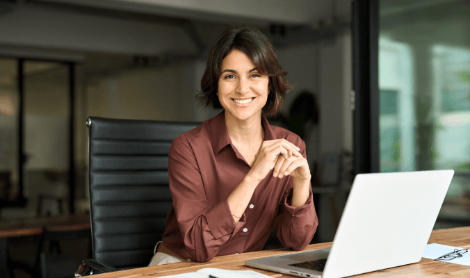 Eine lächelnde Frau sitzt an einem Schreibtisch mit Laptop, symbolisch für den Workshop Frauen & Finanzen.